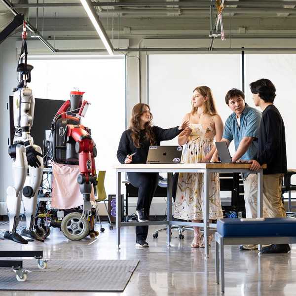 A group of six CWRU students talking in a collaboration space with two robots nearby.